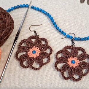 May include: A pair of crocheted flower earrings made with brown and peach yarn. The earrings are hanging from silver hooks. The background is a white surface with a blue beaded necklace.