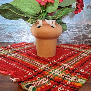 May include: A terracotta flower pot with a white rim and multiple holes for stems. The pot is filled with a bouquet of red flowers with green leaves. The pot is sitting on a red and white patterned table runner.