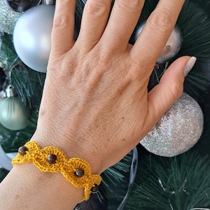 May include: Mustard yellow crochet bracelet with brown beads. The bracelet is made of a woven pattern and is worn on a wrist. The background includes Christmas tree ornaments.