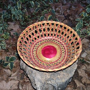Puede incluir: Una cesta tejida con un patrón rojo y marrón. La cesta es redonda y tiene un fondo plano. Está sentada sobre un tocón de árbol.