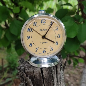 Może przedstawiać: Srebrny budzik z białą tarczą i niebieskimi akcentami. Na tarczy zegara widnieje napis "SLAVA" i "II jewels". Zegar jest ustawiony na godzinę 1:00 i stoi na drewnianym pniu.