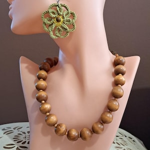 Op de afbeelding: Een groene gehaakte bloemeoorring met een geel centrum, een bruine houten kralenketting en twee garenrollen, een groene en een gele.