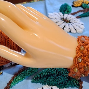 May include: A handmade orange bracelet with a braided design, adorned with small, round, light brown beads. The bracelet is displayed on a mannequin hand, with a ball of orange thread in the background. The bracelet is a piece of jewelry.