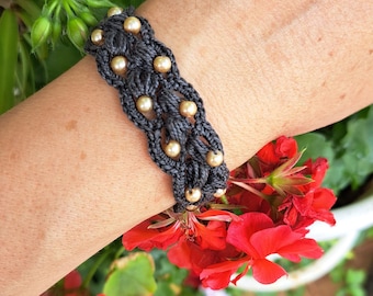 Puede incluir: Un brazalete de macramé gris con cuentas doradas. El brazalete está en la muñeca de una persona y el fondo es una imagen borrosa de flores rojas.