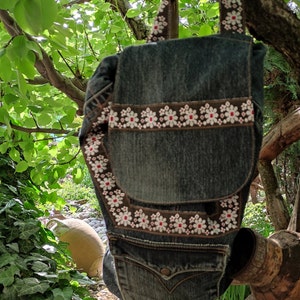 Puede incluir: Un bolso de mezclilla con bordado de flores blancas y rojas. El bolso está colgado de una rama de árbol.