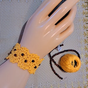May include: A yellow crocheted bracelet with dark brown beads is displayed on a mannequin hand. A ball of yellow thread and a strand of brown beads are also visible. The bracelet has a floral design.