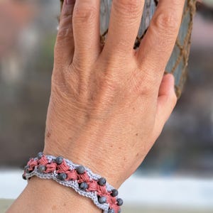 Puede incluir: Un brazalete de crochet gris y rosa con cuentas grises. El brazalete está en la muñeca de una persona.