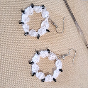 Peut inclure: Une paire de boucles d'oreilles en crochet avec des perles noires. Les boucles d'oreilles sont blanches et ont une forme circulaire.