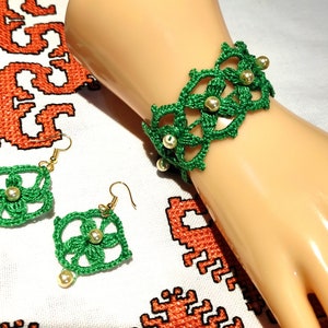 Könnte beinhalten: Ein grünes gehäkeltes Armband und Ohrringe mit weißen Perlenakzenten. Das Armband befindet sich an einem Mannequin-Handgelenk und die Ohrringe auf einem weißen Stoffhintergrund.