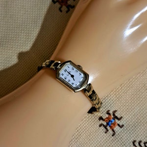 May include: A gold-toned wristwatch with a white face and black Roman numeral hour markers. The watch has a rectangular face and a gold-toned metal band.