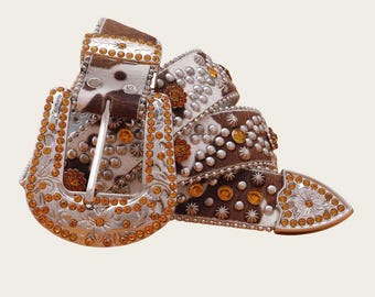 Puede incluir: Un cinturón decorativo con hebilla y detalles plateados. El cinturón presenta un patrón de piel de vaca con parches marrones y blancos, adornado con tachuelas plateadas y pedrería naranja. La hebilla y la punta también están adornadas con pedrería naranja.
