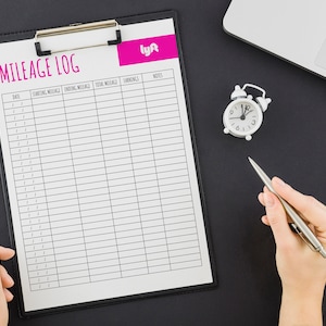 May include: A mileage log sheet with a pink Lyft logo in the top right corner. The sheet is on a black clipboard with a white alarm clock and a silver pen on the black surface.