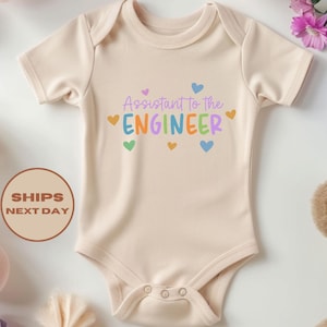 May include: A beige baby bodysuit featuring the text "Assistant to the Engineer" in colourful letters and hearts. The bodysuit is surrounded by decorative paper balls and a teddy bear.