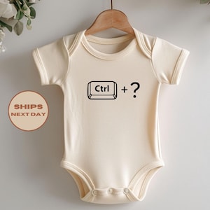 May include: A cream-colored short-sleeve baby bodysuit featuring a black graphic print on the chest. The graphic displays a keyboard control key icon labeled Ctrl followed by a plus sign and a question mark symbol. The garment hangs on a hanger.