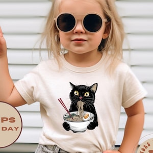 May include: A toddler wearing a beige t-shirt with a black cat eating ramen noodles. The cat is holding chopsticks and there is an egg in the bowl. The toddler is wearing black sunglasses and denim jeans.