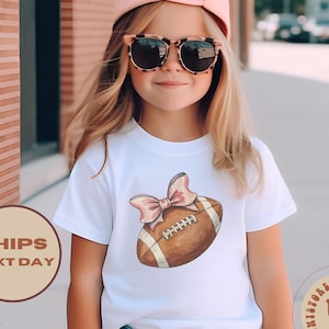 May include: White t-shirt with a brown and white football with a pink bow on top. The shirt has the text "UniStore Kids US" on the bottom right corner.