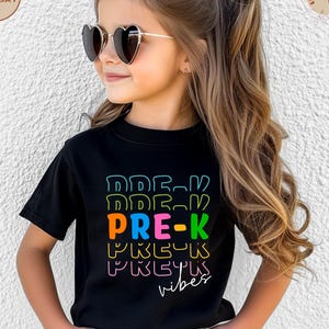 Peut inclure: Un t-shirt noir avec l'inscription "PRE-K vibes" en lettres colorées et soulignées. Le t-shirt est porté par une jeune personne portant des lunettes de soleil en forme de cœur.