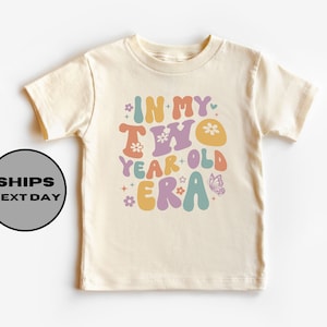 May include: A light beige toddler t-shirt with the text "In My Two Year Old Era" in colorful, retro-style lettering. The text is surrounded by small flowers and a butterfly.