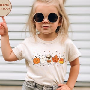 Op de afbeelding: Een crèmekleurig T-shirt met korte mouwen en een grafische print van een lachende pompoen, spook, koffiebeker, herfstblad en maissnoepje. Het ontwerp bevat kleine steraccenten en wordt gecombineerd met een lichtgrijze spijkerbroek.