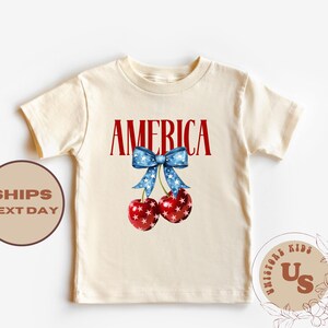 Op de afbeelding: Cr&egrave;mekleurig T-shirt met het woord "AMERICA" in het rood, een blauwe strik versierd met witte sterren en twee kersen. De kersen zijn rood met witte sterpatronen. Een patriottisch ontwerp voor kinderen.