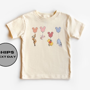 May include: A cream colored t-shirt with a graphic of Winnie the Pooh, Tigger, Piglet, and Eeyore holding onto Mickey Mouse shaped balloons. The balloons are in shades of pink, blue, and yellow.