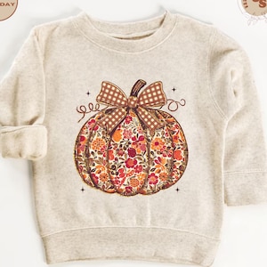 May include: A beige sweatshirt featuring a floral pumpkin design. The pumpkin is filled with red, orange, and yellow flowers, topped with a brown plaid bow and stem. The design includes decorative swirls and small stars.