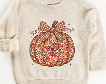 May include: A beige sweatshirt featuring a floral pumpkin design. The pumpkin is filled with red, orange, and yellow flowers, topped with a brown plaid bow and stem. The design includes decorative swirls and small stars.