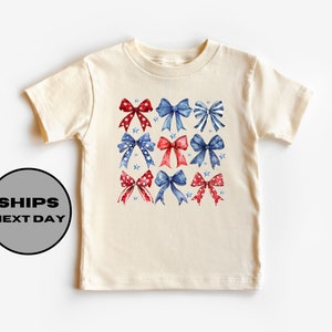 May include: A cream-colored t-shirt with a red, white, and blue bow pattern. The bows are decorated with stars and are arranged in a grid pattern.