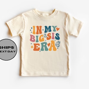 May include: A light beige short-sleeved t-shirt with the text "IN MY BIG SIS ERA" in colorful retro-style lettering with butterflies and stars.