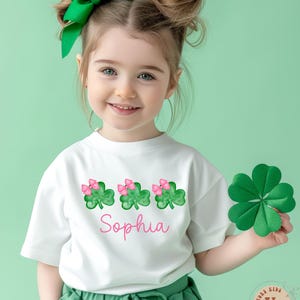 May include: White t-shirt with a green shamrock design and pink bows. The shirt has the name "Sophia" printed on the front.