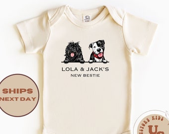 Puede incluir: Un body de bebé de color crema con una ilustración en blanco y negro de dos perros. El texto "Lola & Jack's New Bestie" está impreso en el body.