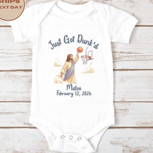 May include: A white short-sleeve baby bodysuit featuring a graphic of a figure dunking a basketball into a hoop. The text reads Just Got Dunk'd, Mateo, and February 12, 2026. The design includes clouds and the figure is wearing a robe.