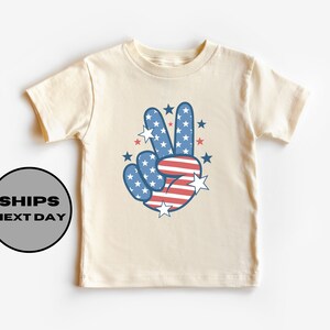 May include: A cream colored t-shirt with a peace sign design. The peace sign is in the colors of the American flag, red, white, and blue. There are stars around the peace sign.