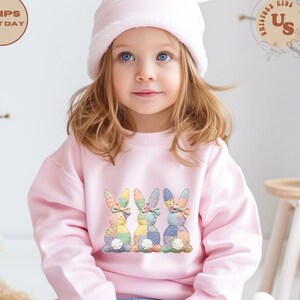 May include: A young child wearing a light pink sweatshirt with a colorful patchwork bunny design. The child is also wearing a matching pink hat with bunny ears. The sweatshirt features three bunnies with patterned ears and bows.