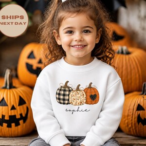 May include: A white long-sleeve sweatshirt featuring a graphic of three pumpkins: one with a black and white buffalo check pattern, one with a floral print, and one orange pumpkin with a black heart. The name Sophie is printed below the pumpkins.