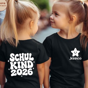 May include: A black short-sleeved t-shirt featuring white graphic text on the back that reads SCHUL KIND 2026 with small star accents. The front of the shirt displays a white smiling star icon above the name Jessica in a white script font.