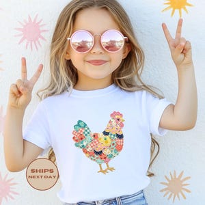 May include: White t-shirt featuring a colorful patchwork chicken design. The chicken has a red comb and yellow feet. The shirt is a classic crew neck style.