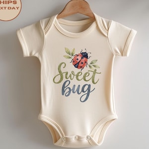 May include: A cream-coloured short-sleeve baby bodysuit featuring a graphic of a red ladybird with green leaves. The text Sweet bug is printed in a cursive font, with Sweet in olive green and bug in a muted blue colour. The garment has a round neckline.