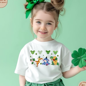 May include: A white t-shirt with a green shamrock and four cartoon characters holding green shamrock balloons. The characters are Winnie the Pooh, Tigger, Piglet, and Eeyore. The shirt is being worn by a young girl with her hair in two buns.