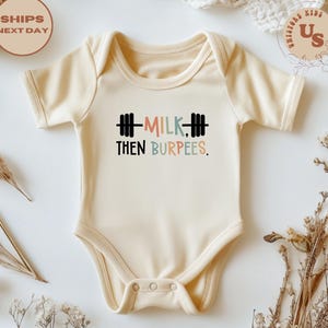 May include: Cream-colored baby onesie with the text "MILK, THEN BURPEES" in black, pink, and blue. The text is accompanied by barbell graphics. The onesie has short sleeves and a snap closure. The background is white with dried flowers.