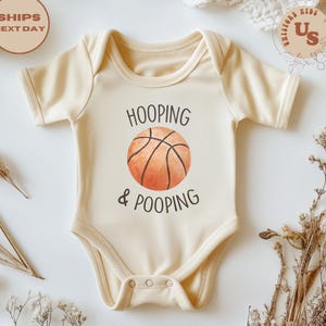 May include: A cream-coloured baby onesie featuring the text "HOOPING & POOPING" and a basketball graphic. The onesie has short sleeves and poppers at the bottom. The design is centered on the front of the garment.
