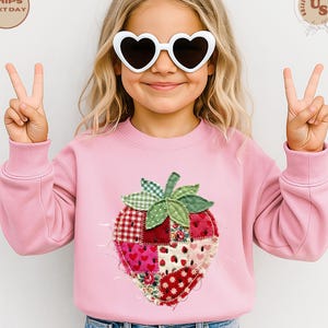 May include: A pink sweatshirt featuring a large, patchwork strawberry design. The strawberry has a green and white checkered leaf, and the patches are red, pink, and white with various patterns. The child is wearing heart-shaped sunglasses.