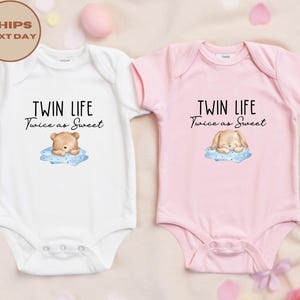 May include: Two baby onesies, one white and one pink, with the text "TWIN LIFE Twice as Sweet." The white onesie features a bear illustration, and the pink one has a bunny illustration. Both illustrations are on a blue cloud.