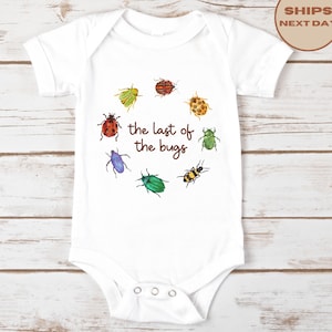 May include: A white short-sleeved bodysuit featuring a circular arrangement of colorful illustrated insects surrounding the brown text "the last of the bugs" printed in the center. The garment has a round neckline and three metal snaps at the bottom.