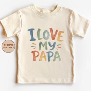 May include: Cream-colored t-shirt with the text "I LOVE MY PAPA" in colorful, watercolor-style letters. The shirt is a short-sleeved, crew neck style, perfect for a child.