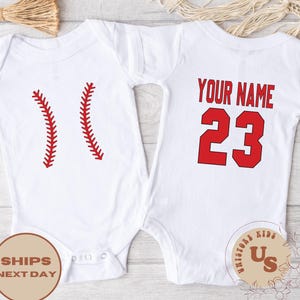 May include: Two white baby onesies with red baseball-themed designs. One onesie has two red baseball stitches, the other has the text "YOUR NAME" and the number "23" in red.