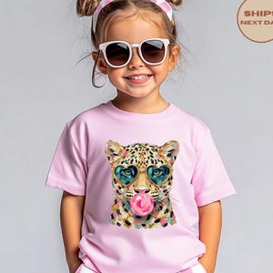 May include: A light pink t-shirt featuring a graphic print of a leopard wearing heart-shaped sunglasses and blowing a pink bubblegum bubble. The leopard has a spotted coat with gold, black, and teal accents, and the shirt has a crew neckline.