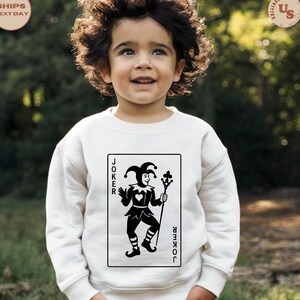 May include: A white sweatshirt featuring a black and white graphic of a joker card. The card design includes a jester figure holding a staff, with the word "JOKER" on either side. The child is smiling.