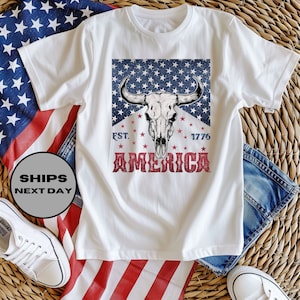 May include: White t-shirt with a red, white, and blue American flag design featuring a skull of a bull with the text "EST. 1776 AMERICA".