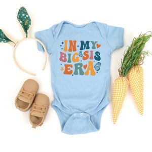 May include: A light blue baby onesie with the text "In My Big Sis Era" in colorful retro font. The onesie is decorated with hearts, butterflies, and stars.